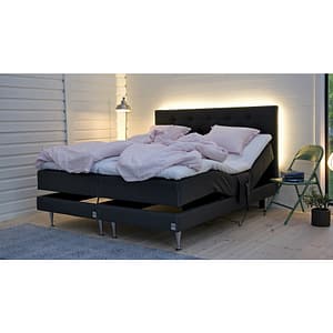 A modern bedroom features the Skanör ställbar “paket” adjustable bed in black, pink bedding, and a tufted headboard with soft backlighting. A green chair with a plant and books sits beside it, under a pendant lamp on the white paneled wall.