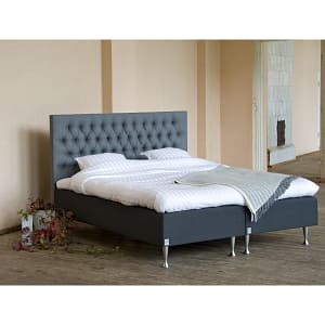 A modern bedroom featuring the Vadstena gray upholstered bed frame with a tufted headboard, white bedding, two pillows, a gray blanket, light walls, wooden flooring, and a potted plant beside the bed.