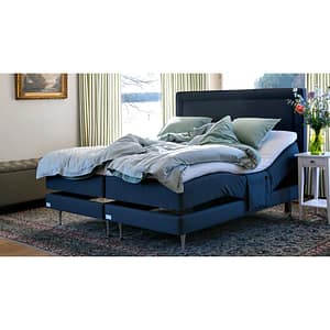 A modern bedroom features the Vaxholm, a large blue adjustable bed with white and light blue bedding, partially elevated. The room includes a floral rug, ottoman, side table with flowers, and large windows with light curtains.