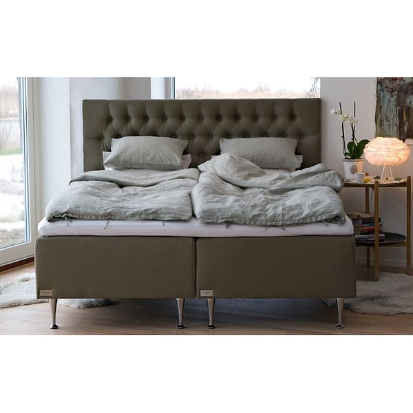 The Exclusive double bed with a tufted headboard, plush bedding, and two pillows stands in a bright room near large windows, beside a side table with a potted orchid and a lit lamp.