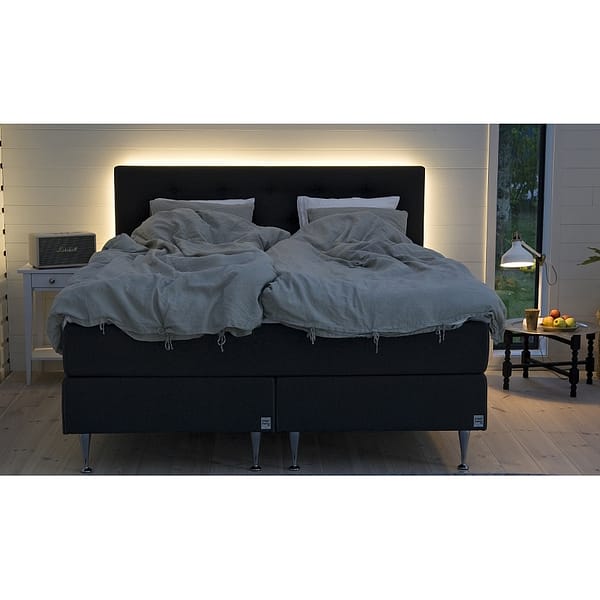 The Skanör “paket” double bed with a dark upholstered headboard stands in a softly lit, cozy bedroom. Gentle light glows behind the headboard, flanked by nightstands topped with lamps and decorative accents on both sides.