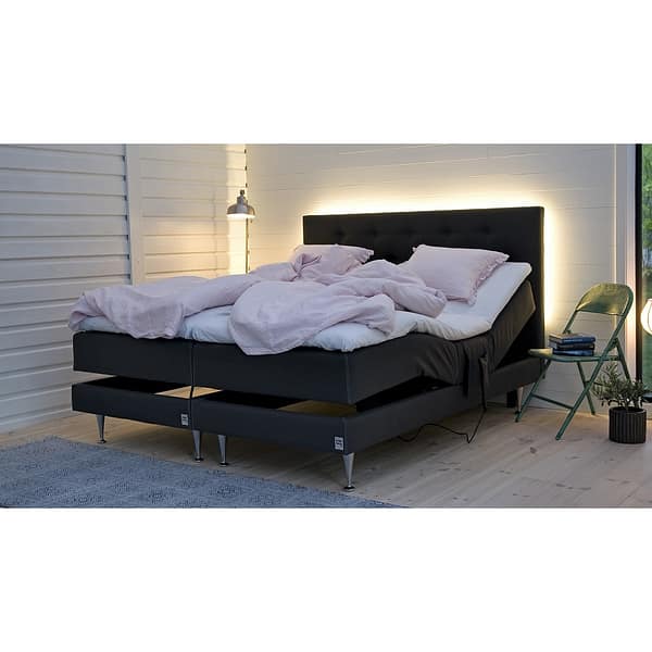 A modern bedroom features the Skanör ställbar “paket” adjustable bed in black, pink bedding, and a tufted headboard with soft backlighting. A green chair with a plant and books sits beside it, under a pendant lamp on the white paneled wall.